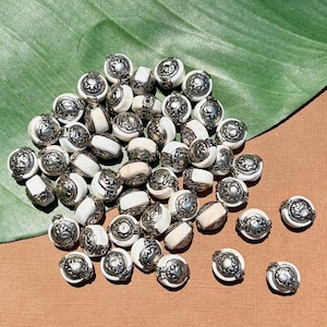 Könnte beinhalten: Eine Ansammlung runder Perlen mit einem weißen, glatten Mittelteil, der von zwei verzierten, strukturierten silberfarbenen Metallkappen flankiert wird. Jede Perle hat ein einheitliches Design mit geprägten Mustern an den Metallenden.