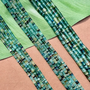May include: Two strands of turquoise blue glass beads, each strand is made up of many small, irregular shaped beads. The beads are arranged in a line and are on a green and brown surface.