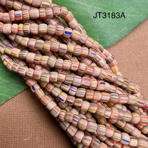 May include: A collection of small, cylindrical beads in a pale pink base color, each featuring horizontal stripes of blue, yellow, orange, and white. The beads are arranged in multiple strands, and the text JT3183A is visible in the upper right corner.
