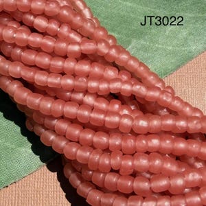 Puede incluir: Una colección de pequeñas cuentas redondas con acabado mate en un color rosa suave y apagado. Las cuentas están ensartadas en varias filas paralelas, creando un grupo denso y texturizado. El texto JT3022 es visible en la esquina superior derecha.