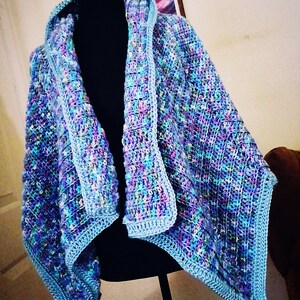 Puede incluir: Un chal tejido a crochet con cuello con capucha en tonos azules, morados y verdes. El chal se muestra en un maniquí negro.