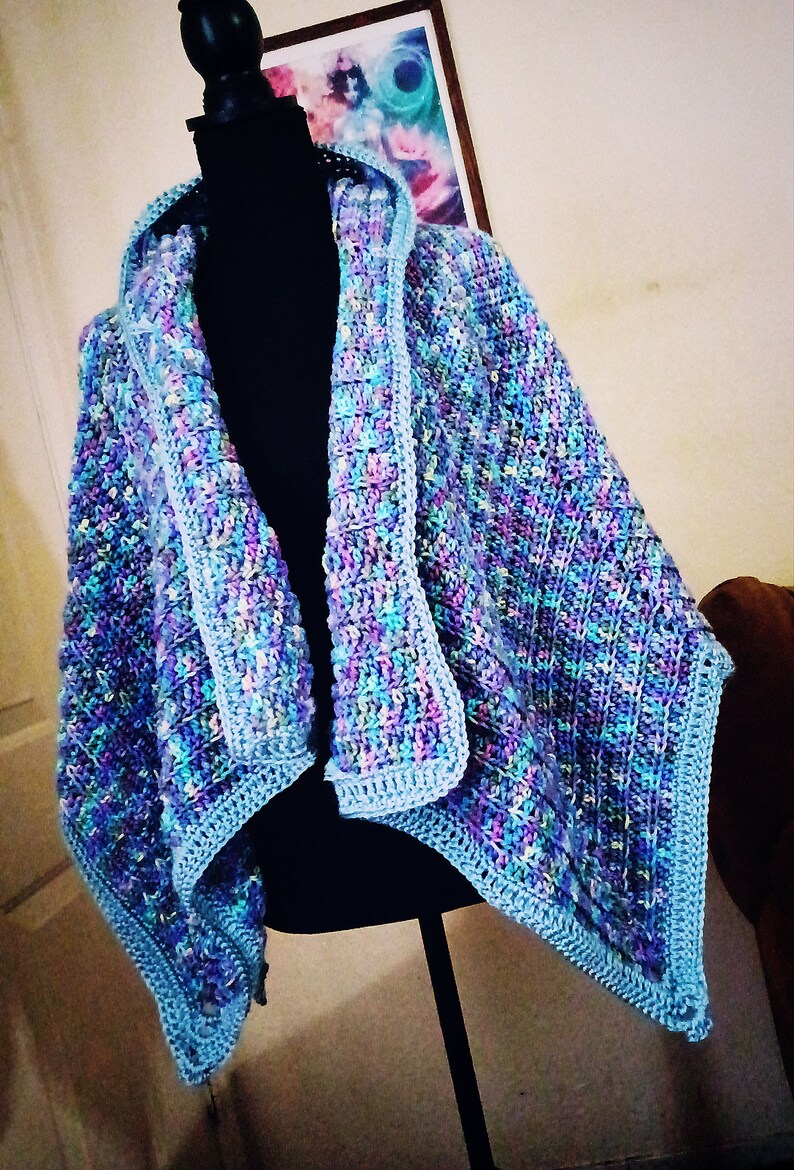 K&ouml;nnte beinhalten: Ein geh&auml;keltes Tuch mit Kapuze in Blaut&ouml;nen, Lila und Gr&uuml;n. Das Tuch wird auf einer schwarzen Schaufensterpuppe pr&auml;sentiert.