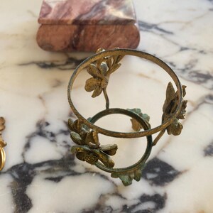 May include: An antique brass-colored decorative stand with floral accents. The stand has two circular rings connected by floral stems. A small, rectangular marble box with a lid is in the background. The marble box has a pink and brown pattern.