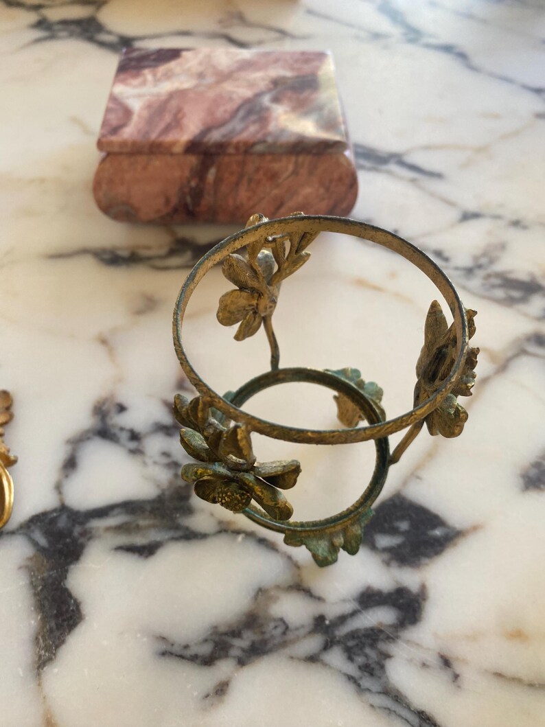 May include: An antique brass-colored decorative stand with floral accents. The stand has two circular rings connected by floral stems. A small, rectangular marble box with a lid is in the background. The marble box has a pink and brown pattern.
