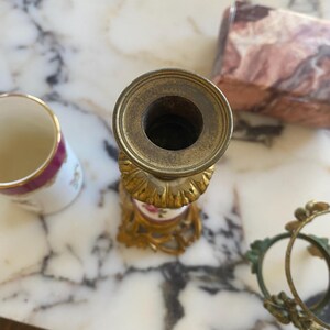 May include: An antique brass candlestick holder with a floral design on a white and pink porcelain base. A small porcelain cup with a pink rim and floral design is next to the candlestick. A decorative gold frame is also visible.