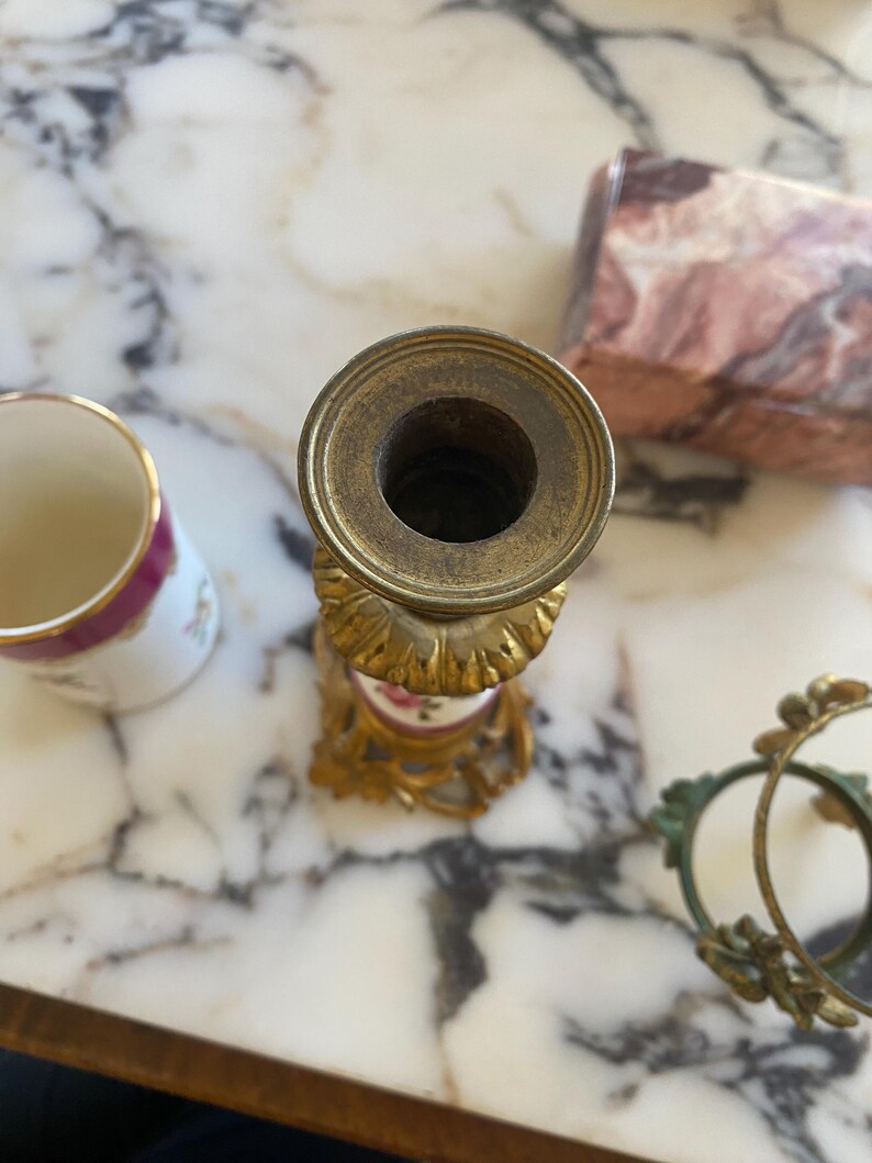 May include: An antique brass candlestick holder with a floral design on a white and pink porcelain base. A small porcelain cup with a pink rim and floral design is next to the candlestick. A decorative gold frame is also visible.
