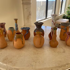 May include: A collection of handmade ceramic vases in various shapes and sizes. The vases feature a warm, earthy color palette with dripped glaze details in shades of yellow, green, blue, and red. The vases are displayed on a round, marble-like table.