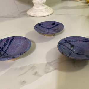 May include: Three small, purple ceramic dishes with black painted designs. The dishes are on a white countertop.