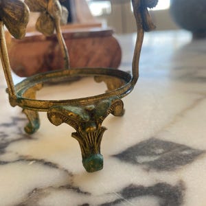 May include: An antique gold-colored metal egg holder with a decorative floral design. The holder has three ornate legs and a circular top, with a weathered, aged appearance. The metal has hints of green patina.