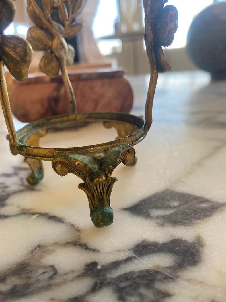 May include: An antique gold-colored metal egg holder with a decorative floral design. The holder has three ornate legs and a circular top, with a weathered, aged appearance. The metal has hints of green patina.