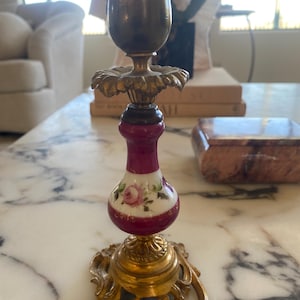 May include: A vintage brass and porcelain candlestick holder. The base and top are brass, while the center features a maroon and white porcelain section with a painted rose design. The candlestick holder has an ornate, decorative design.