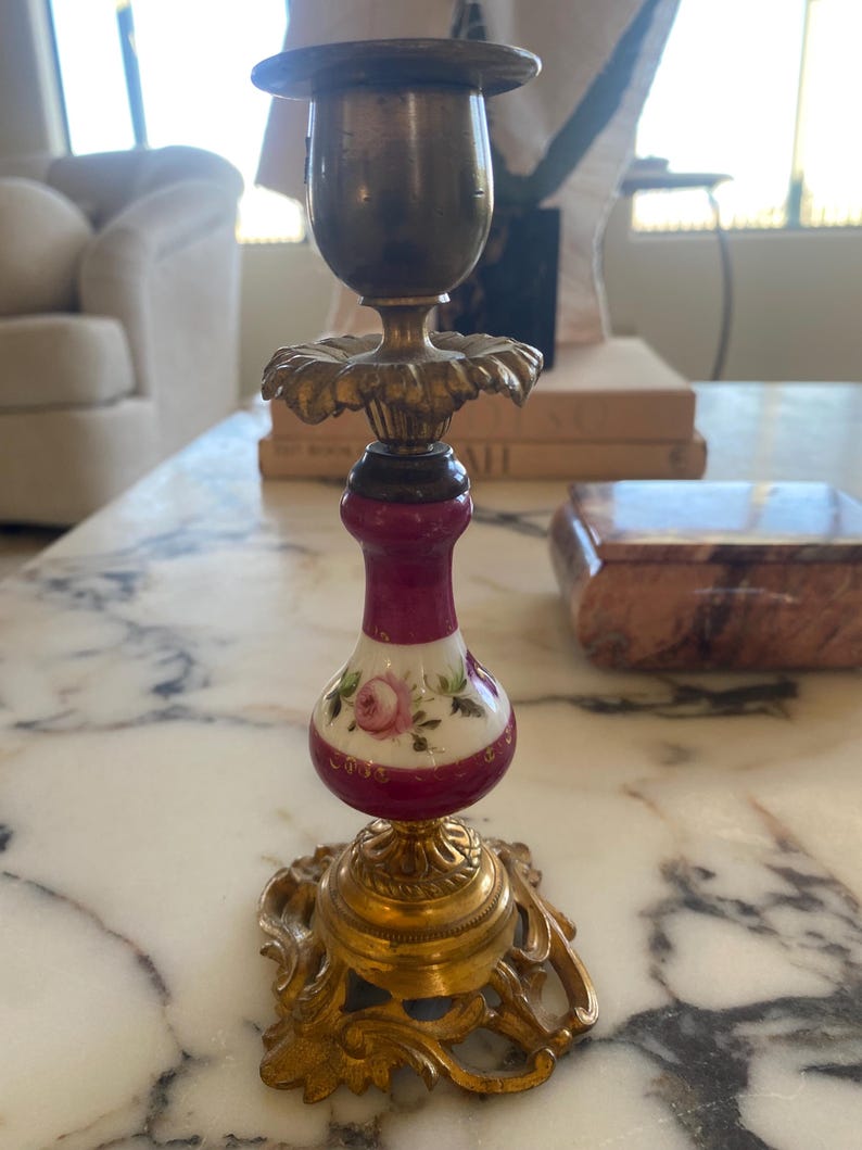 May include: A vintage brass and porcelain candlestick holder. The base and top are brass, while the center features a maroon and white porcelain section with a painted rose design. The candlestick holder has an ornate, decorative design.