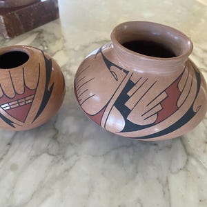 May include: Two handcrafted pottery vessels with earth-tone colors and geometric designs. The smaller pot is round with a wide opening, while the larger one has a wider body and a narrower opening. Both pots feature black, red, and white patterns.