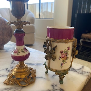 May include: Two antique decorative items on a marble surface. One is a brass and porcelain candlestick with a burgundy and floral design. The other is a small vase with a burgundy rim, floral design, and brass accents.