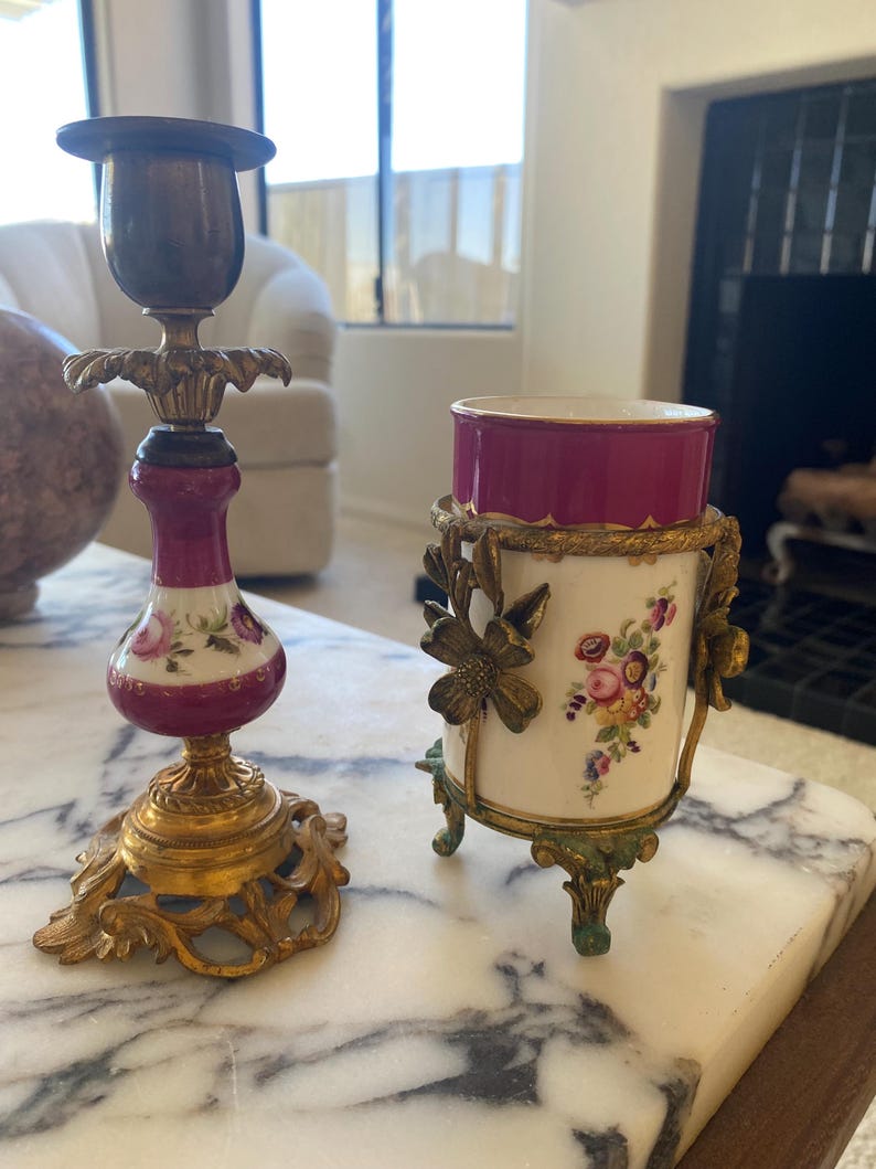 May include: Two antique decorative items on a marble surface. One is a brass and porcelain candlestick with a burgundy and floral design. The other is a small vase with a burgundy rim, floral design, and brass accents.