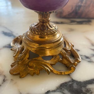 May include: A decorative, ornate gold-colored stand with a purple and white egg-shaped object on top. The stand has a detailed, scrolled base and a pedestal. The egg-shaped object has a purple base and a white top.