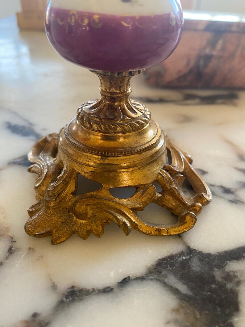 May include: A decorative, ornate gold-colored stand with a purple and white egg-shaped object on top. The stand has a detailed, scrolled base and a pedestal. The egg-shaped object has a purple base and a white top.