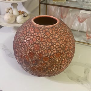 May include: A round, textured vase with a reddish-brown exterior and a dark brown interior. The vase has a unique, bumpy surface pattern. Decorative items are visible in the background, suggesting a home decor theme.