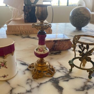 May include: An assortment of antique decorative items, including a brass and porcelain candlestick with floral accents, a small brass stand, and a porcelain cup with a burgundy rim and floral design. The items are arranged on a marble surface.