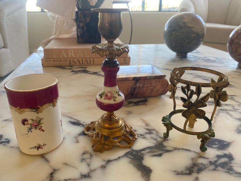 May include: An assortment of antique decorative items, including a brass and porcelain candlestick with floral accents, a small brass stand, and a porcelain cup with a burgundy rim and floral design. The items are arranged on a marble surface.