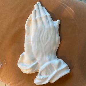 May include: A white plaster sculpture of two hands clasped in prayer. The hands are detailed with visible knuckles and wrinkles, and the sculpture has a smooth, matte finish. The piece is likely a religious or decorative item.