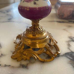 May include: A vintage perfume bottle with a white top adorned with pink roses and green leaves. The bottle has a deep purple midsection and sits on a gold-colored ornate base. The base has a detailed, scrolled design.