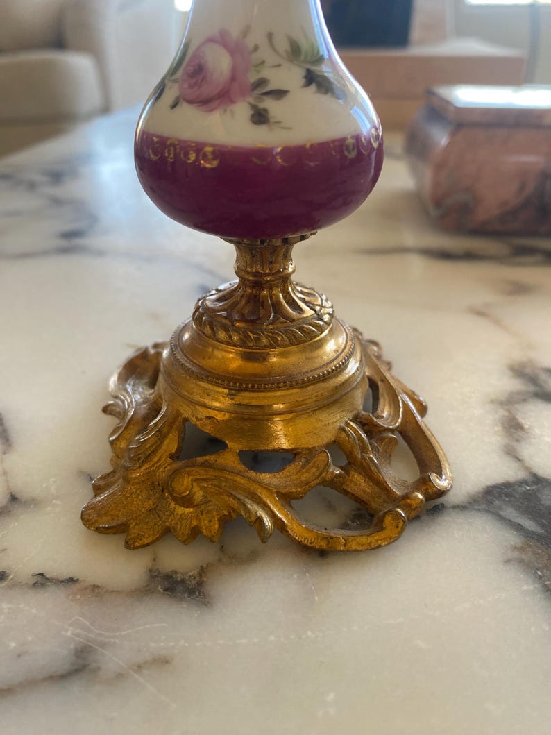 May include: A vintage perfume bottle with a white top adorned with pink roses and green leaves. The bottle has a deep purple midsection and sits on a gold-colored ornate base. The base has a detailed, scrolled design.