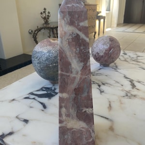 May include: A tall, four-sided obelisk carved from polished stone featuring a variegated pattern of deep reddish-brown, muted grey, and creamy white veins. The stone sculpture has a pointed pyramidal top and a flat, square base.