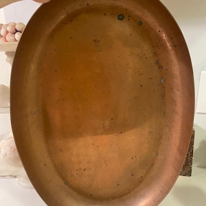 May include: Oval copper tray with two handle cutouts. The tray has a warm, reddish-brown hue with visible patina and minor imperfections. The tray is held by a hand, suggesting its size and portability. This vintage tray is suitable for serving or display.