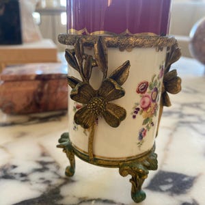 May include: A decorative porcelain vase with a burgundy top and floral design. The vase is held by a gold-colored metal frame with floral accents and rests on three ornate feet. The vase is a beautiful antique.