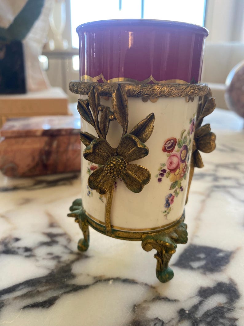 May include: A decorative porcelain vase with a burgundy top and floral design. The vase is held by a gold-colored metal frame with floral accents and rests on three ornate feet. The vase is a beautiful antique.