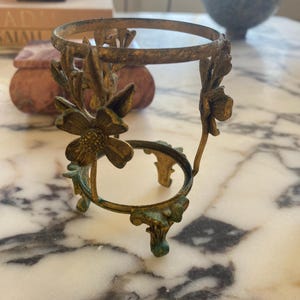 May include: Ornate, antique-style brass sphere stand with floral accents. The stand has a circular top and bottom, connected by curved supports with flower details. The brass has a weathered, aged appearance with hints of green patina.