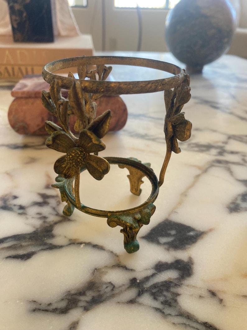May include: Ornate, antique-style brass sphere stand with floral accents. The stand has a circular top and bottom, connected by curved supports with flower details. The brass has a weathered, aged appearance with hints of green patina.