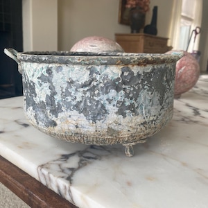 May include: A weathered, round metal planter with a distressed, textured finish in shades of gray, white, and teal. The planter has small feet and handles. The planter is sitting on a marble surface.