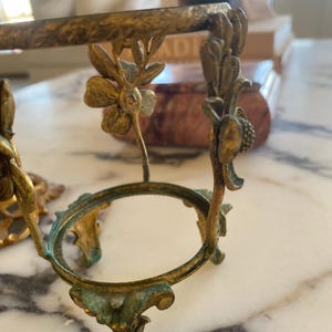 May include: Ornate, antique brass-toned egg holder with floral and leaf details. The holder features a circular base and decorative supports, showcasing a vintage aesthetic. The brass has a slight patina, adding to its aged charm.