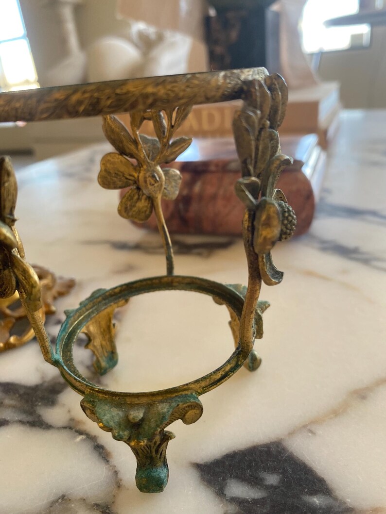 May include: Ornate, antique brass-toned egg holder with floral and leaf details. The holder features a circular base and decorative supports, showcasing a vintage aesthetic. The brass has a slight patina, adding to its aged charm.