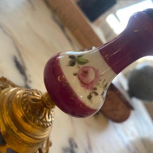 May include: A decorative, ornate gold-colored metal fixture with a floral design. The fixture is attached to a handle with a maroon and white porcelain knob. The knob features a painted pink rose and gold accents.