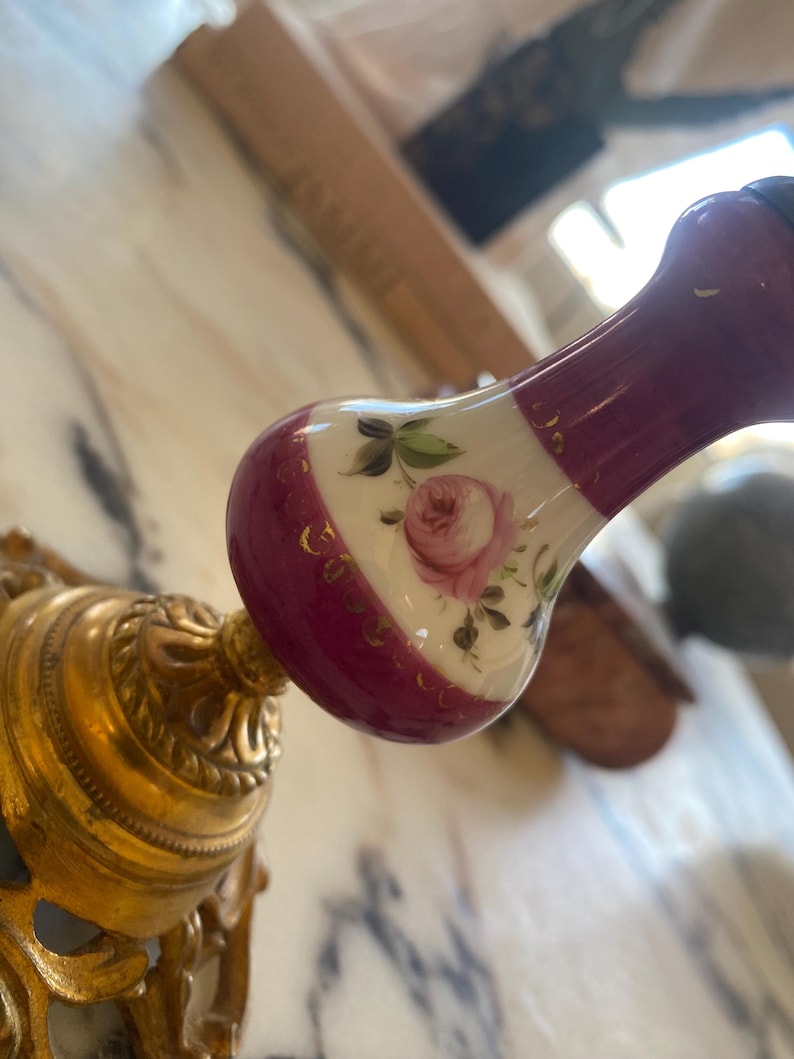 May include: A decorative, ornate gold-colored metal fixture with a floral design. The fixture is attached to a handle with a maroon and white porcelain knob. The knob features a painted pink rose and gold accents.