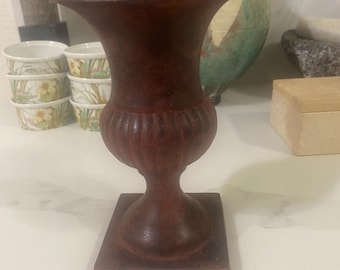 May include: A dark brown, urn-shaped decorative vase with a square base. The vase has a fluted body and a wide, flared top. Other items in the background include small bowls with floral designs and a large, blue and green vase.