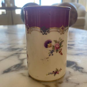 May include: A cylindrical, antique porcelain cup with a deep purple rim and gold trim. The cup features a cream-colored body adorned with floral bouquets in shades of purple, pink, and blue. The cup is approximately 4 inches tall.