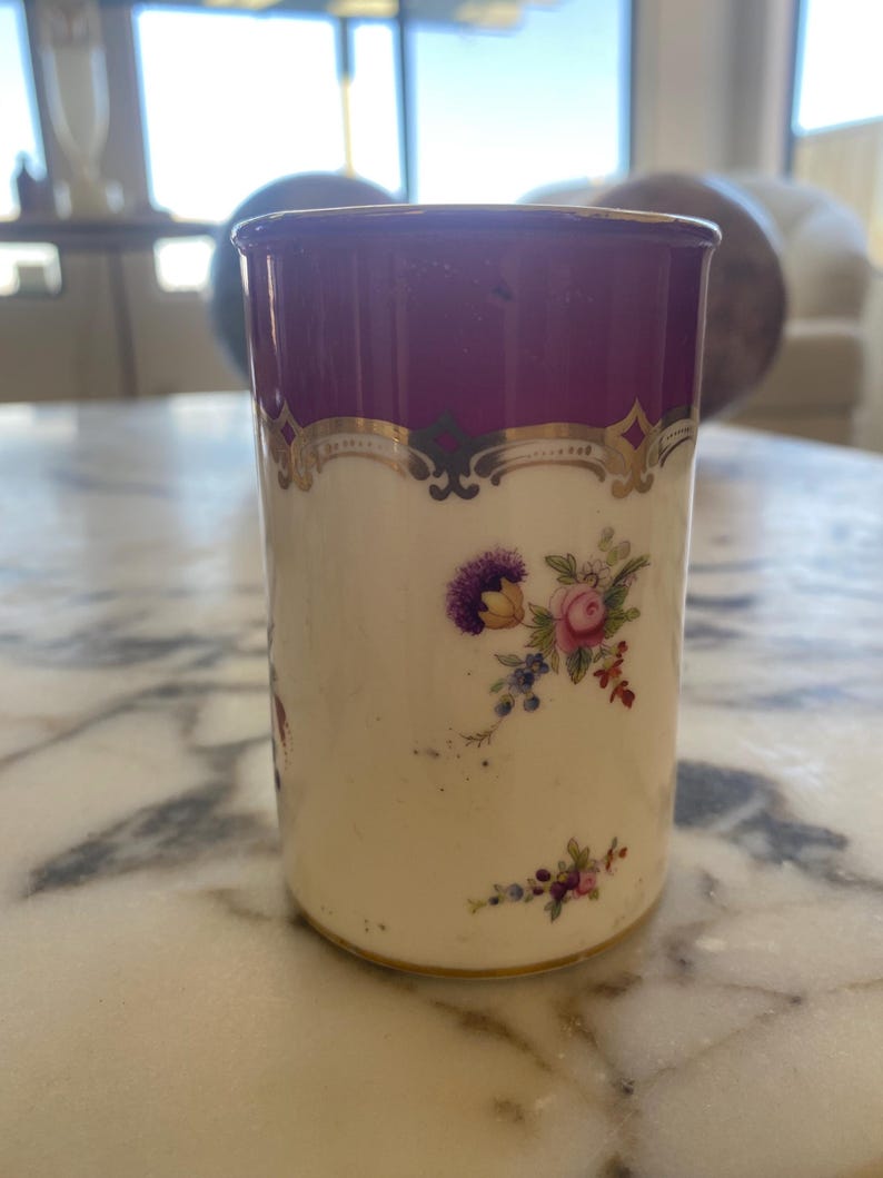 May include: A cylindrical, antique porcelain cup with a deep purple rim and gold trim. The cup features a cream-colored body adorned with floral bouquets in shades of purple, pink, and blue. The cup is approximately 4 inches tall.