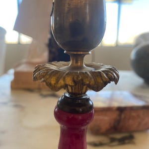May include: Close-up of an antique brass candlestick holder. The holder features a gold-colored floral design and a burgundy-colored base with floral patterns. The top portion is a tarnished brass color.