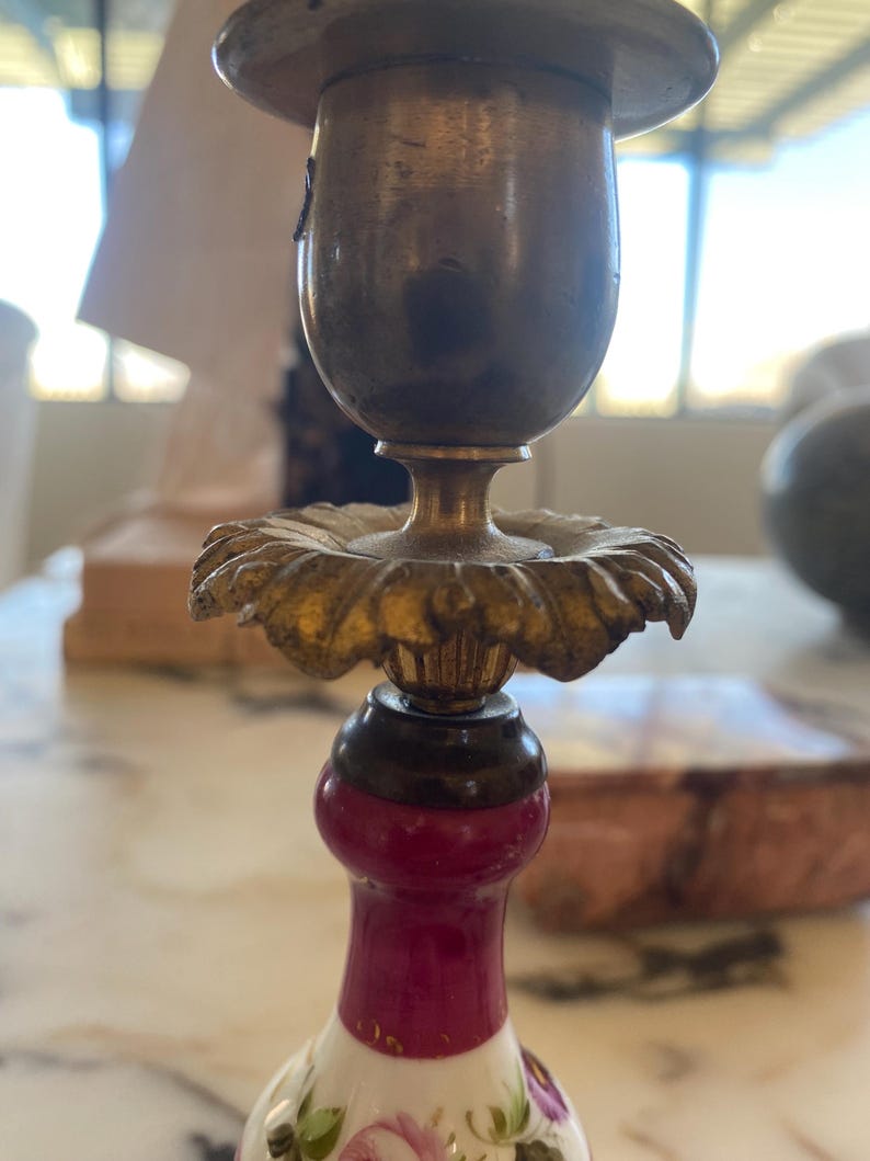 May include: Close-up of an antique brass candlestick holder. The holder features a gold-colored floral design and a burgundy-colored base with floral patterns. The top portion is a tarnished brass color.