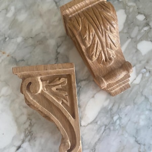 May include: Two light brown wooden corbels featuring intricate carved leaf patterns and scrollwork details. These architectural elements have a natural wood grain finish and a flat top surface, showcasing detailed craftsmanship in the wood carving.