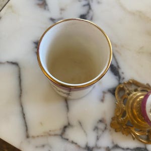 May include: A small, white ceramic cup with a gold rim. The cup has a simple design with a maroon floral pattern. The cup is sitting on a marble surface. A gold-colored decorative object is in the background.
