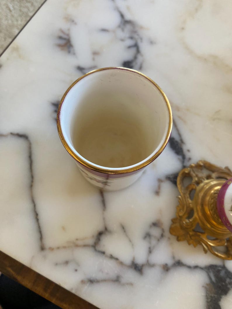 May include: A small, white ceramic cup with a gold rim. The cup has a simple design with a maroon floral pattern. The cup is sitting on a marble surface. A gold-colored decorative object is in the background.