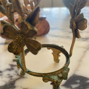 May include: Ornate, antique-style metal stand with a circular top and floral accents. The stand has a weathered, aged appearance with a mix of gold and verdigris green tones. The design features delicate flower and leaf details.