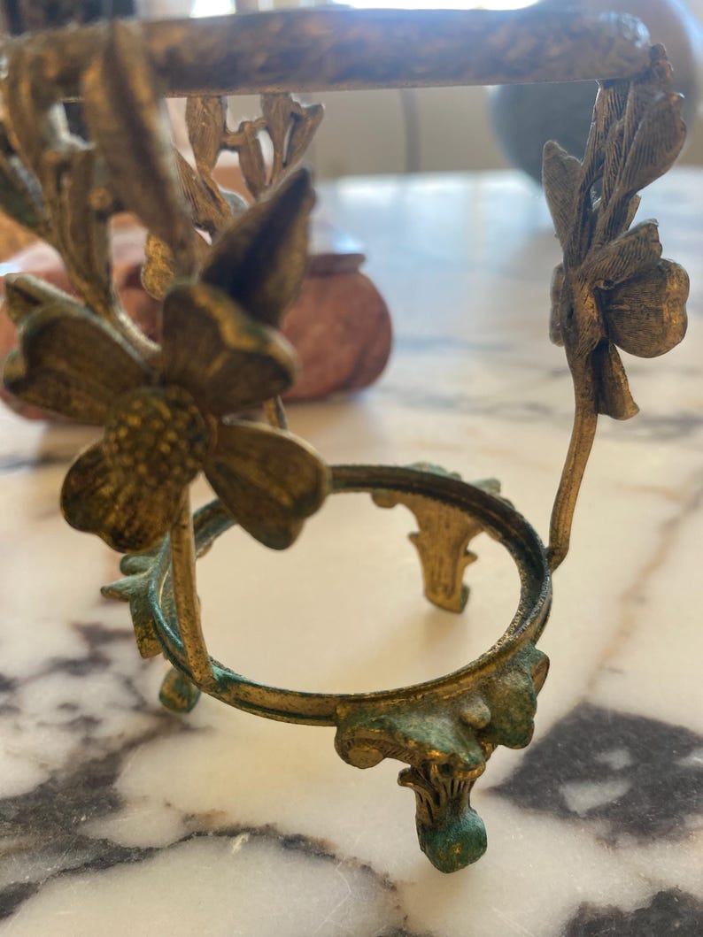 May include: Ornate, antique-style metal stand with a circular top and floral accents. The stand has a weathered, aged appearance with a mix of gold and verdigris green tones. The design features delicate flower and leaf details.