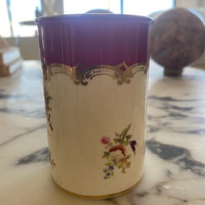 May include: A cylindrical porcelain container with a cream-colored body and a deep purple rim. The container is adorned with gold filigree and delicate floral patterns in shades of pink, blue, and yellow.