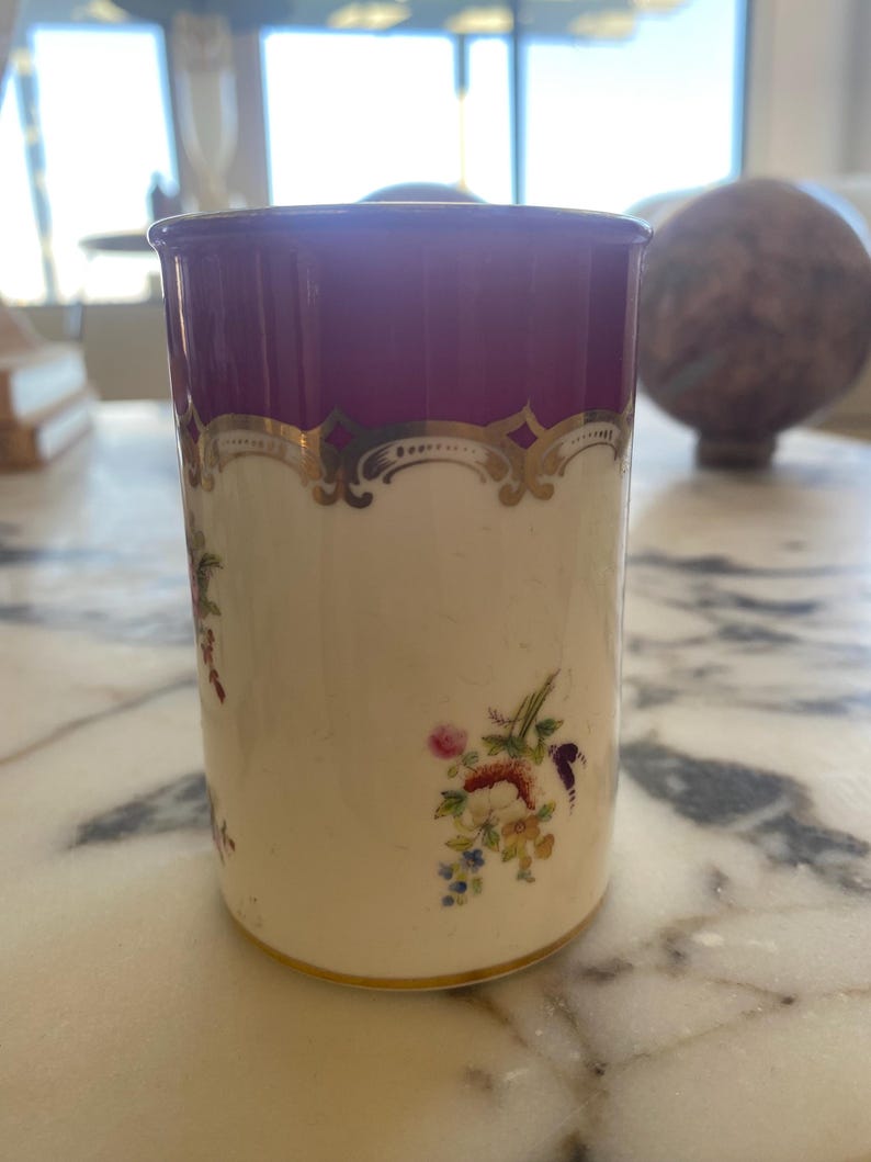 May include: A cylindrical porcelain container with a cream-colored body and a deep purple rim. The container is adorned with gold filigree and delicate floral patterns in shades of pink, blue, and yellow.